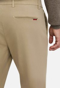 Pantalones beige con una textura suave, que cuentan con un bolsillo trasero con un acento rojo. La cinturilla está equipada con trabillas para ajustar.