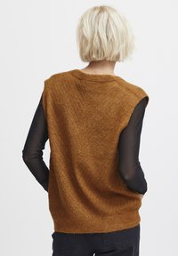 Brauner, gestrickter, ärmelloser Pullover mit geripptem Muster und Zickzack-Design, kombiniert mit einem transparenten schwarzen Langarmoberteil darunter.