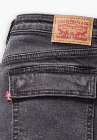 Jeans de mezclilla gris con una etiqueta de cuero marrón que presenta gráficos rojos y negros en la cintura y un pequeño parche rojo en el bolsillo trasero.