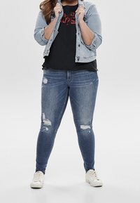 Vaalean siniset farkut, joissa on kulutuspintaa, yhdistetty mustaan graafiseen t-paitaan ja vaaleansiniseen farkkutakkiin. Valkoiset tennarit viimeistelevät asun.