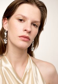 Boucles d'oreilles argentées avec un design fluide et sculptural. Le mannequin porte un haut halter en satin beige lisse avec un léger éclat.