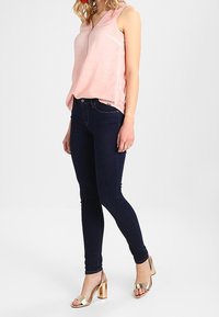 Haut sans manches rose avec un décolleté en V et des fentes sur les côtés, associé à un jean skinny bleu foncé et des sandales à talons dorées métalliques. Look simple et décontracté.