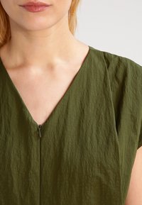 Vestido verde oliva con una tela texturizada, que presenta un escote en V y un pequeño detalle de cremallera en la parte delantera para el cierre.