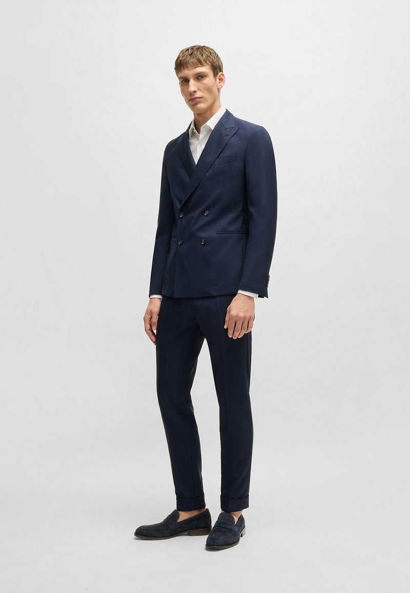 Costume croisé bleu marine avec revers en pointe, associé à une chemise blanche et des mocassins foncés. Tissu lisse avec une coupe ajustée.