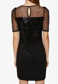 Černé šaty s krátkými průhlednými rukávy, zdobené flitrovaným zadním dílem s postupným vzorem. Detaily ve tvaru klíčové dírky na zadní části výstřihu.