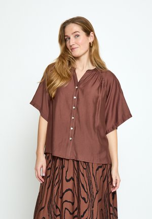 Kobieta o długich blond włosach, ubrana w luźną brązową koszulę zapinaną na guziki i brązową spódnicę z czarnym abstrakcyjnym wzorem, stojąca na białym tle.