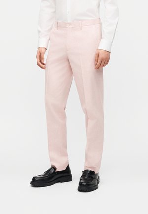 Muškarac u svijetloružičastim šivenim hlačama uparenom s bijelom košuljom i crnim kožnim mokasinkama, stoji na bijeloj pozadini.