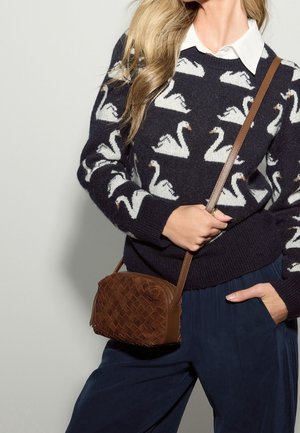 Femme portant un pull marine avec des motifs de cygnes blancs, une chemise blanche à col, un pantalon marine, et tenant un sac bandoulière tressé marron.