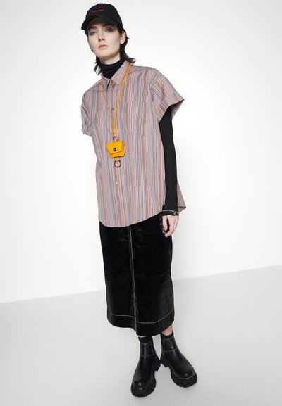 Camisa de mangas curtas às riscas em cores pastel, saia preta brilhante, botas pretas robustas e um acessório em forma de câmara amarela pendurado numa corrente.