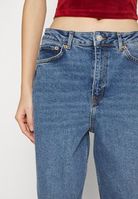 Calças de ganga azul com cintura alta, design de cinco bolsos e costuras contrastantes. O tecido tem uma textura suave e ligeira descoloração.