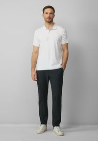 Witte polo met een gestructureerd patroon, gecombineerd met donkere broeken en witte sneakers, staande tegen een effen grijze achtergrond.