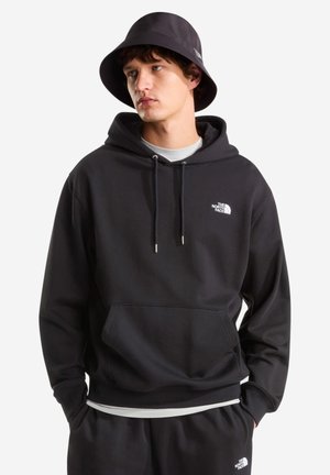 Jonge man draagt een zwarte The North Face hoodie, bijpassende bucket hat en zwarte broek, handen in de zakken, kijkt iets opzij.