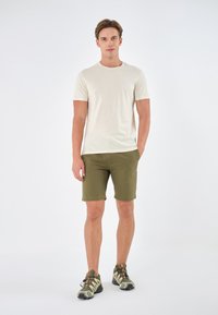 Beige t-skjorte med korte ermer kombinert med olivengrønne shorts, komplettert av kamuflasjemønstrede joggesko med svarte såler og snøringer.