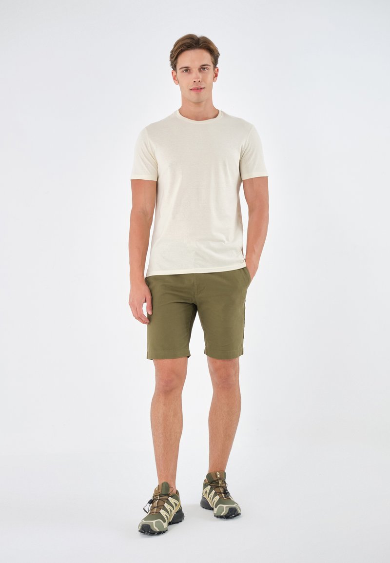 Beige t-skjorte med korte ermer kombinert med olivengrønne shorts, komplettert av kamuflasjemønstrede joggesko med svarte såler og snøringer.