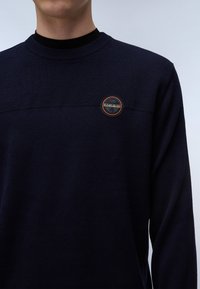 Marineblauwe gebreide trui met een gestructureerd bovenstuk en een ronde logopatch op de borst. Lange mouwen en ronde hals.