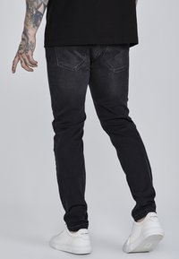Black denim jeans with a slim fit, featuring back pockets and subtle fading, paired with white sneakers on a light background.