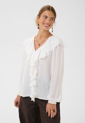 Glimlachende vrouw die een witte blouse met lange mouwen en ruches, een donkere broek en gouden sieraden draagt, staand tegen een effen lichte achtergrond.