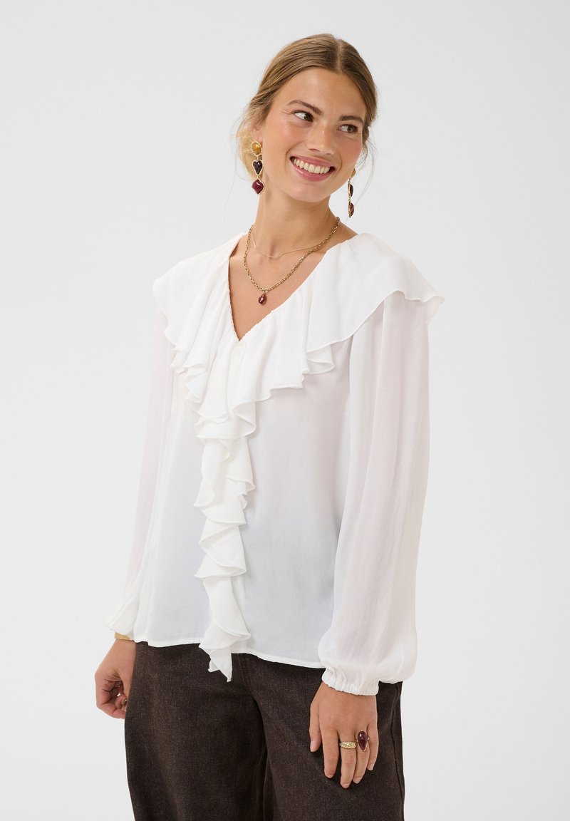Femme souriante portant un chemisier blanc à manches longues avec des volants, un pantalon foncé et des bijoux en or, debout devant un fond uni clair.