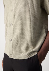 Lichtgrijze, geribbelde gebreide cardigan met knopen aan de voorkant en een ronde zoom. Bevat textiel met structuur en contrasterende lichte knopen. Draagt over donkere broeken.