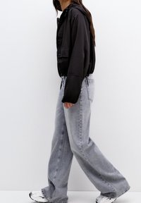 Giacca nera con cappuccio e zip frontale, abbinata a jeans a gamba larga grigi chiari. Il modello indossa sneakers bianche. Un look semplice e casual.