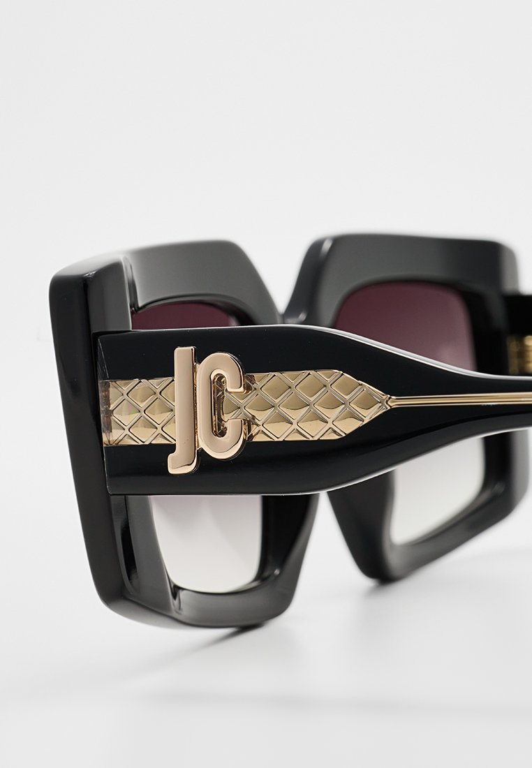 Schwarze übergroße Sonnenbrille mit glanzvoller Oberfläche. Eigenschaften umfassen ein Logo in Goldton und eine strukturierte goldene Akzentuierung an den Bügeln. Die Gläser verlaufen von pink zu klar.