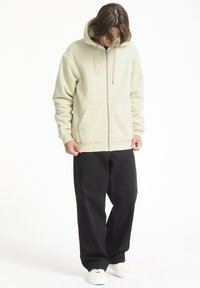 Sudadera beige con cremallera, bolsillos frontales y capucha ajustable con cordón, combinada con pantalones negros de corte holgado y zapatillas ligeras.