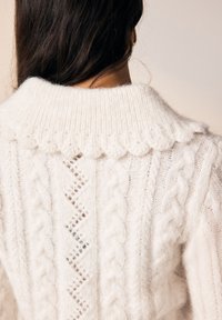 Kremasto obarvan pleten pulover z okroglim ovratnikom z valovitimi robovi, teksturo kablaste pletenine in vzorci v obliki diamantov na hrbtni strani.