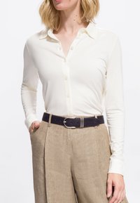 Kvinde iført en hvid langærmet knapshirt, der er stoppet ned i beige højtaljede bukser med et sort bælte, hånd i lommen, mod en hvid baggrund.