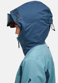 Blauwe en groene sneeuwjas met een grote capuchon, een rits en een logo. Bevat een skibril die op het voorhoofd is bevestigd, wat het ontwerp van de band benadrukt.