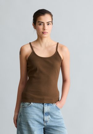 Jeune femme avec un débardeur côtelé marron et un jean bleu clair, debout devant un fond gris uni avec une main dans la poche.