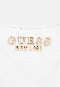 Vitt texturerat material med präglade blomsterdekorationer. Guldmetalliskt "GUESS EST 1981"-logotyp visas framträdande.