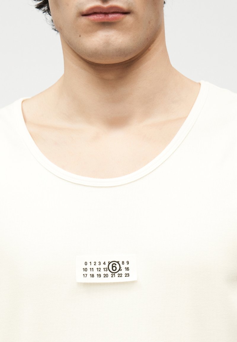 Primo piano di una persona che indossa una canottiera bianca a coste con una piccola etichetta rettangolare che mostra i numeri da 0 a 23, con il numero 6 cerchiato.