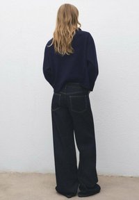 Donna bionda in piedi con le spalle alla macchina fotografica, che indossa jeans scuri larghi e un maglione blu navy, di fronte a un semplice muro grigio chiaro.