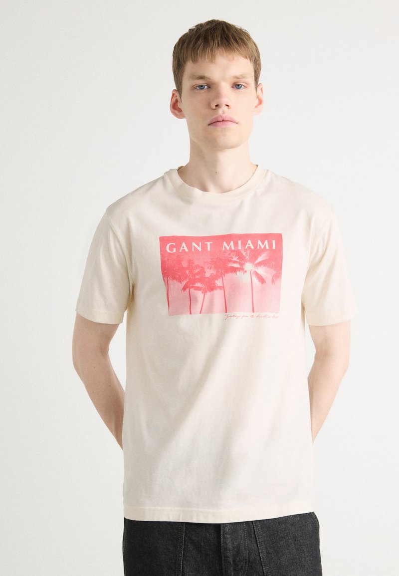 Krémové tričko s krátkým rukávem, které má růžový grafický potisk palem a text "GANT MIAMI". Vyrobeno z jemného bavlněného materiálu.