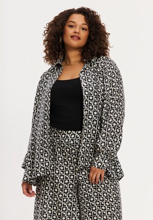 Kvinde med krøllet hår iført en sort top, sort-hvidt åbent skjortemønster med geometrisk design og matchende bukser med vide ben.