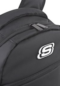 Mochila negra fabricada con nylon duradero, que cuenta con un bolsillo con cremallera, correas acolchadas y un prominente logo blanco en la parte frontal.