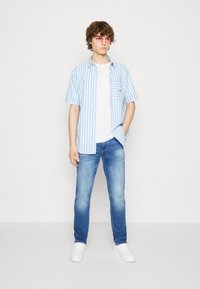 Camicia a maniche corte blu e bianca a righe sopra una t-shirt bianca, abbinata a jeans chiari strappati e sneakers bianche.
