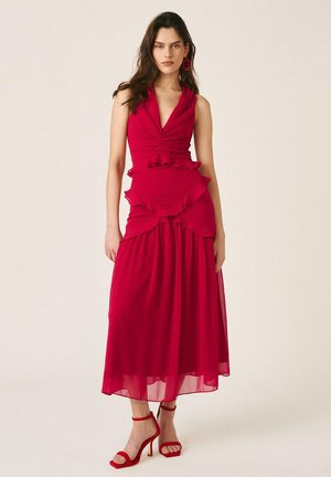 Mujer con vestido rojo sin mangas con volantes en capas en la cintura y las caderas, combinado con sandalias de tacón rojo y pendientes a juego.