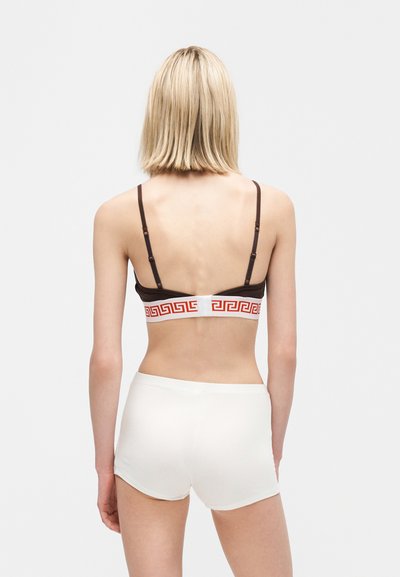 Γυναίκα με κοντά ξανθά μαλλιά που φοράει καφέ σουτιέν με λευκή και κόκκινη ταινία με μοτίβο ελληνικού κλειδιού και λευκά σορτς με ψηλή μέση, που φαίνεται από πίσω.