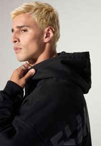 Black hooded sweatshirt with a soft texture, featuring a rounded neckline and a large graphic logo in a darker shade on the back.
