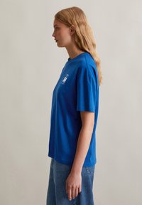 Blaues Baumwoll-T-Shirt mit kurzen Ärmeln und rundem Halsausschnitt; verfügt über ein weißes Druckdesign auf der Brust. Kombiniert mit hellblauen Jeans.