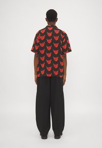 Uomo in piedi di spalle, con una camicia nera a maniche corte con motivo di cuori diavolo rossi e pantaloni larghi neri.