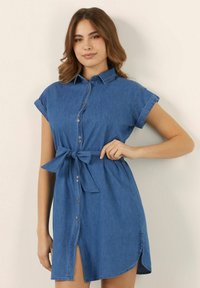 Sininen farkkumekko, jossa on lyhyet käännetyt hihat, napillinen etuosa ja sidottava vyö. Epäsymmetrinen helma, jossa on sivureikä.