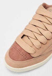 Sneaker in camoscio beige con lacci spessi, area della punta perforata e una sezione in tan scuro a contrasto. Suola in gomma bianca con bordi texturizzati.