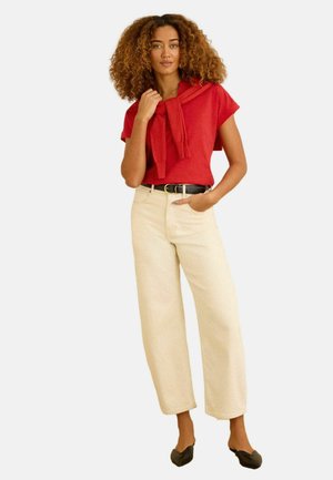 Žena s kudrnatými vlasy v červeném krátkém tričku, béžových širokých kalhotách, černém opasku, černých balerínách a červeném svetru přehozeném přes ramena.