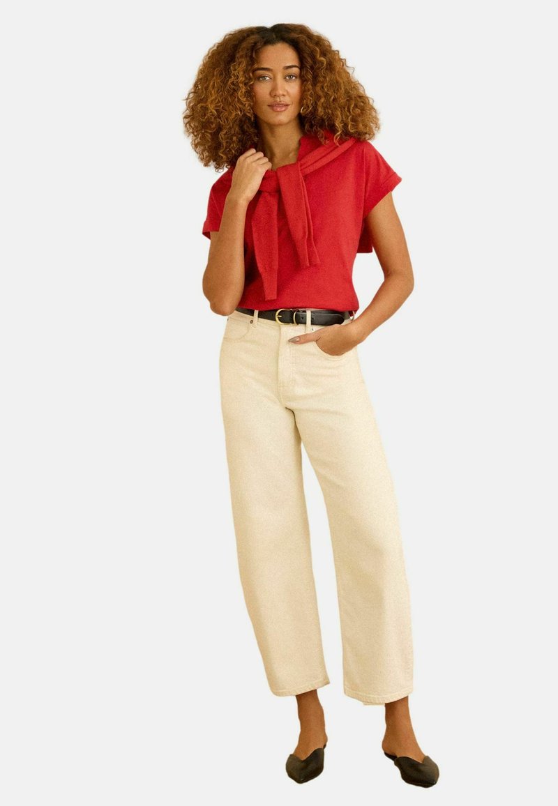 Göndör hajú nő, vörös rövid ujjú felsőt, bézs bőszárú nadrágot, fekete övet, fekete balerinacipőt visel, és a vállára terített piros pulóvert.