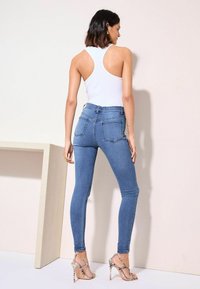 Mėlynos aukštai sukarpytos liesos džinsai su lygiu paviršiumi ir užpakalinėmis kišenėmis, derinami su balta tank topu ir sandalais su aukštu kulnu.