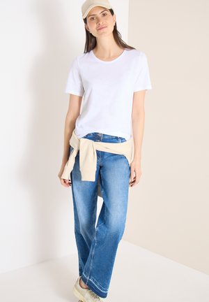 Unge kvinde iført beige kasket, hvid t-shirt, blå jeans, beige sweatshirt bundet omkring taljen og sneakers, stående mod en ensfarvet baggrund.