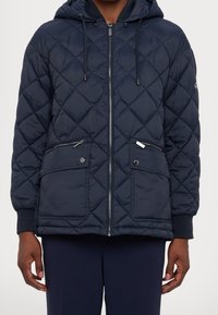 Marineblå quiltet dunjakke med hætte, lynlås, to forlommer, ribkant ved ærmerne og en glat, let tekstur.