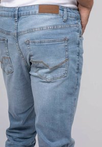 Personne portant un jean en denim bleu clair avec des coutures visibles, des poches arrière et une étiquette de marque en cuir brun sur la taille.